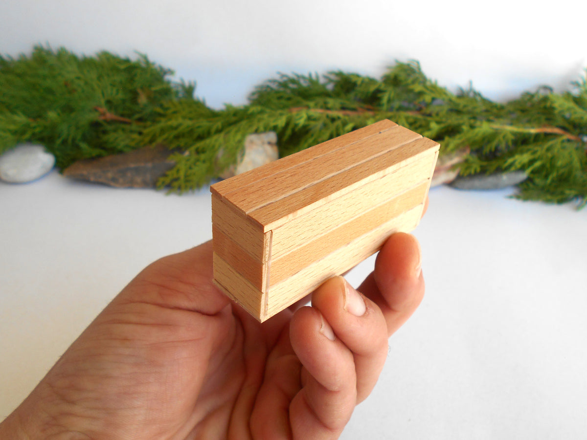 A hand holding a small rectangular wooden box with a herringbone pattern of light and medium wood tones, set against a soft-focus background of green pine branches and white stones.