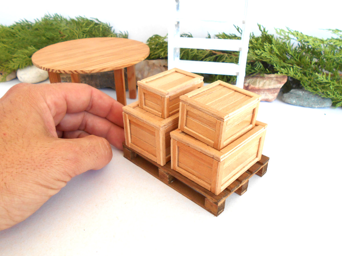 A miniature wooden crate set on a small wooden pallet, with a hand holding it for scale, against a white tabletop with miniature garden props in the background.