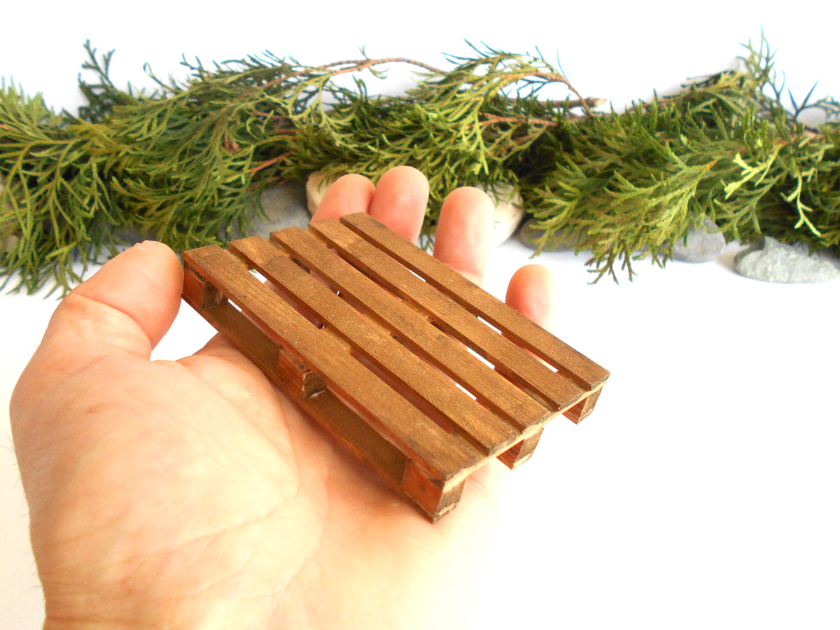 A miniature wooden slat tray or miniature pallet, held in a hand to show its small scale, with a green garland and white background.