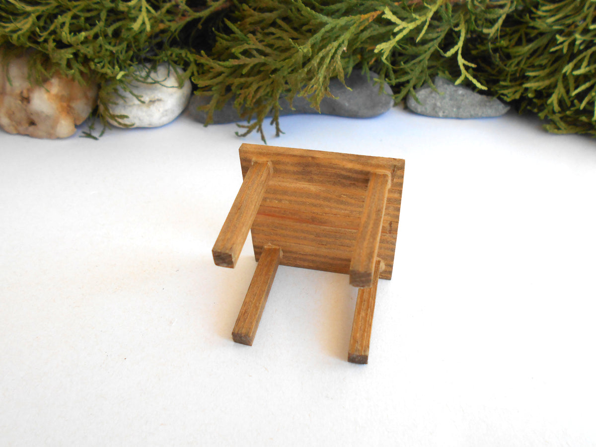 A miniature wooden stool or small chair with a warm medium-brown finish, photographed on a white surface with decorative greenery and stones in the background.