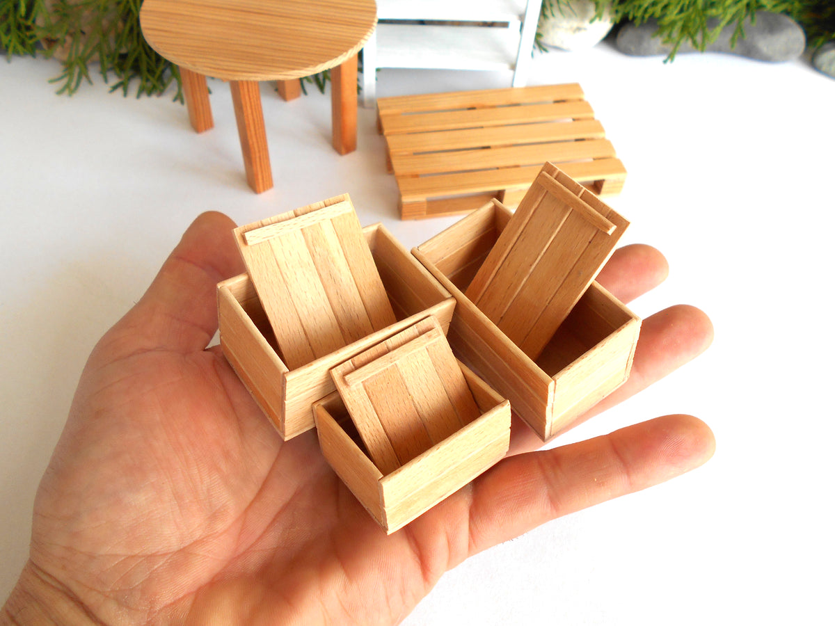 A hand holding several miniature light wood crates, with more matching furniture pieces blurred in the background on a white surface.