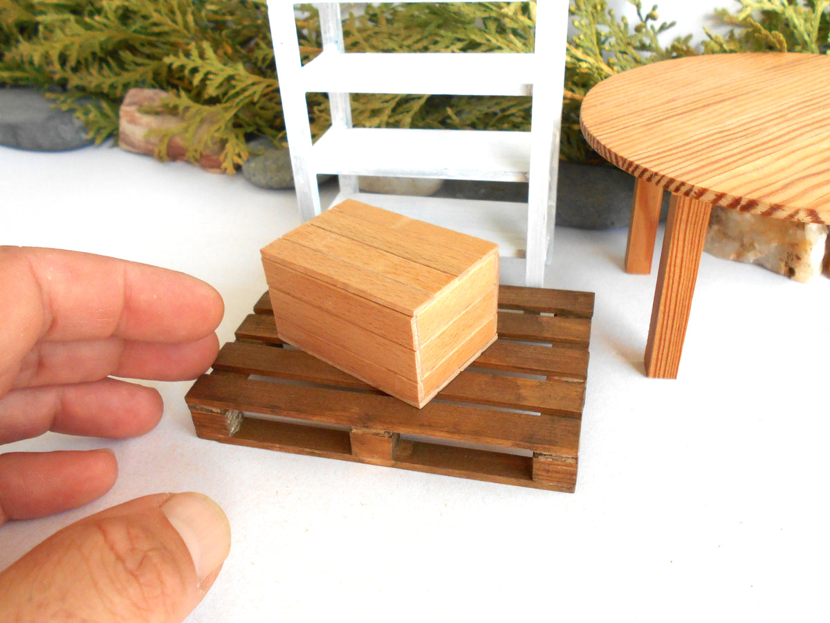 A miniature wooden box placed on a small wooden pallet, with a human hand holding it for scale, set against a miniature diorama background with a white chair and round wooden table.