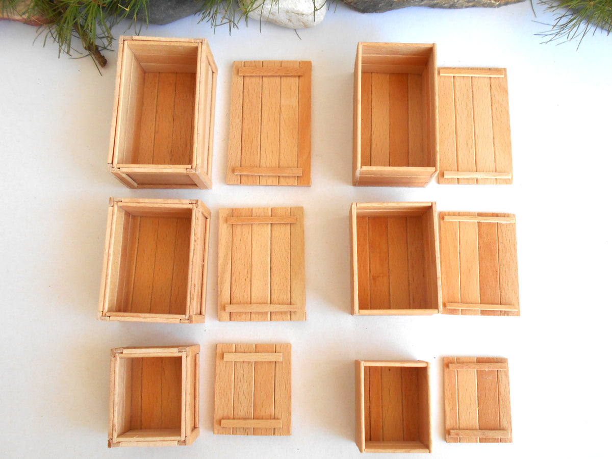 Nine miniature light wood crates and boxes arranged in a grid on a white surface, with small green pine branches visible at the top edge.