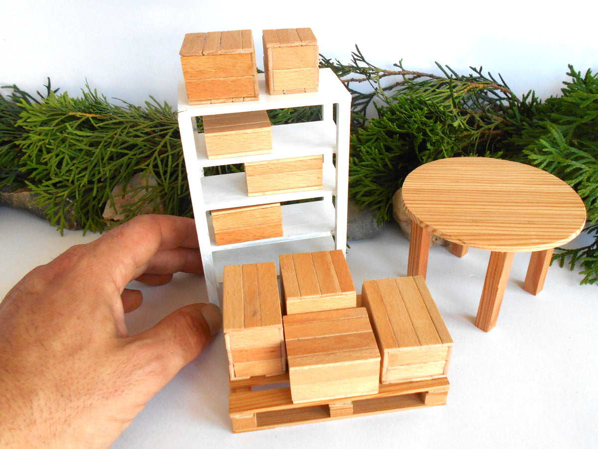 A miniature wooden toy furniture set including a white shelving unit, a round light-wood table, and matching wooden boxes and pallets, shown with a human hand adjusting one of the pieces against a white background with green pine branches.