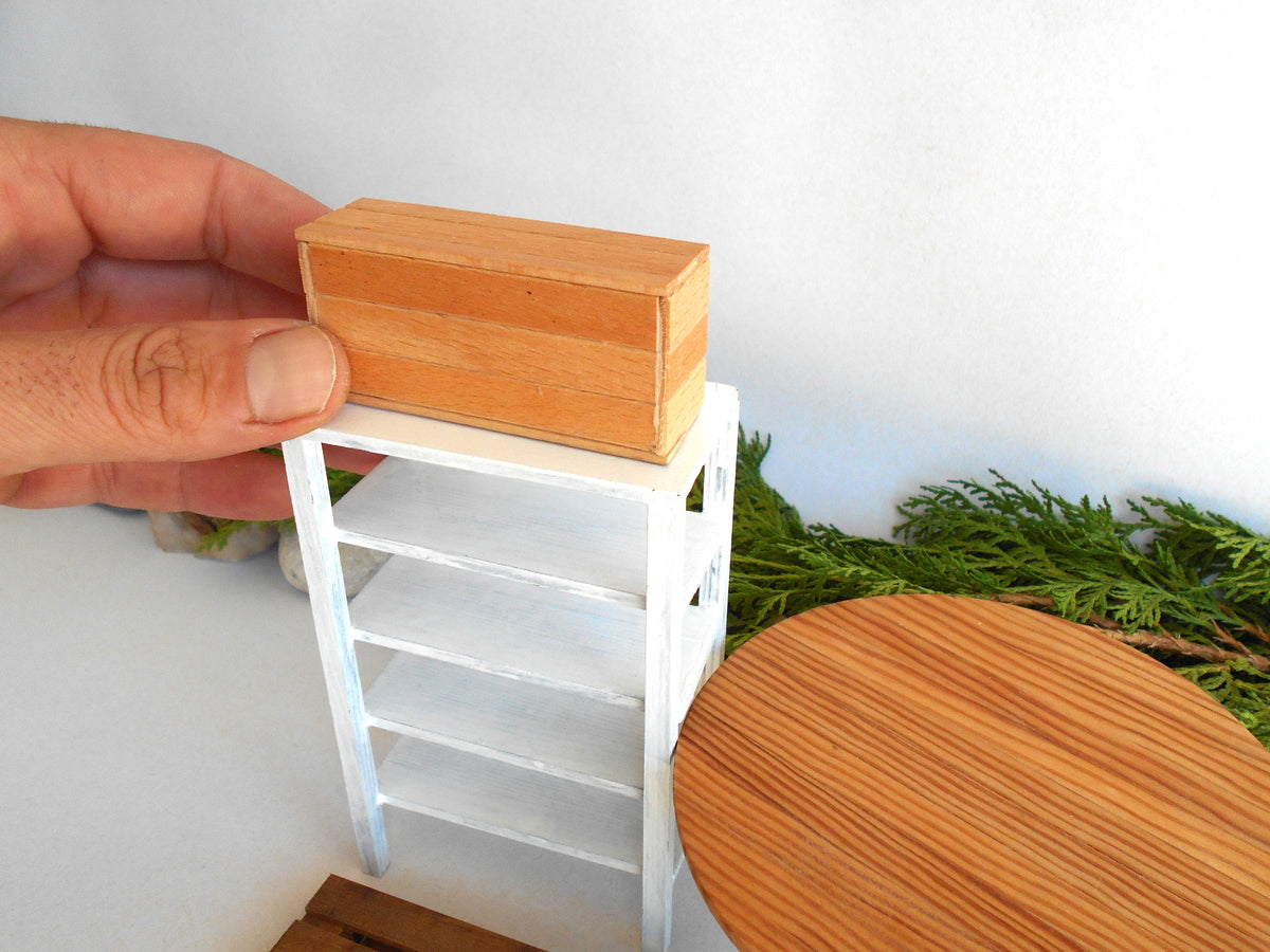 A miniature wooden storage box with a light brown wood grain finish is being held by a hand on top of a small white miniature shelf. The background features a white wall, green foliage, and a miniature round wooden table.