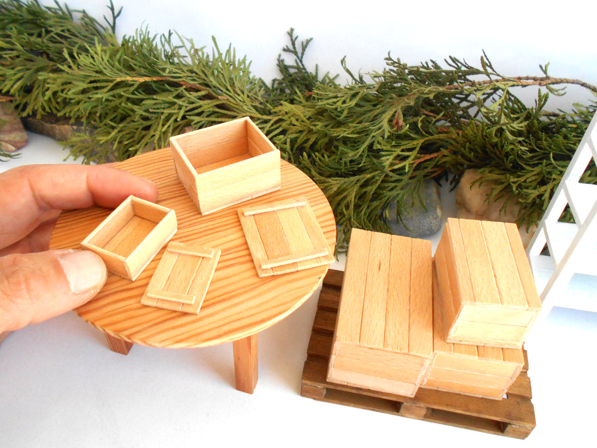 A hand holds a miniature round wooden table with small wooden boxes and lids, while additional small wooden crates and blocks sit on a miniature pallet in a dollhouse-scale setting.