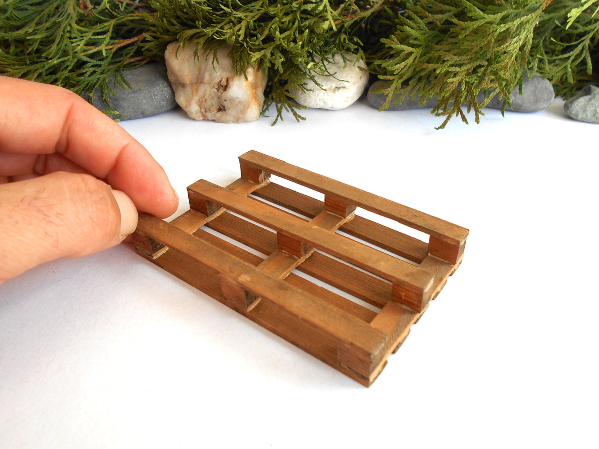 A miniature wooden pallet model being held by a hand, set against a white background with decorative greenery and stones.