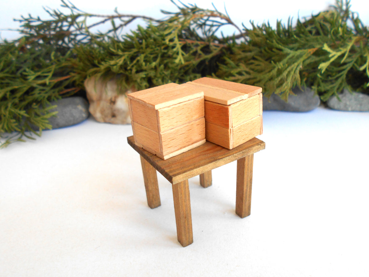 A miniature wooden toy table with two small stacked wooden boxes, photographed on a white surface with blurred green foliage and stones in the background.