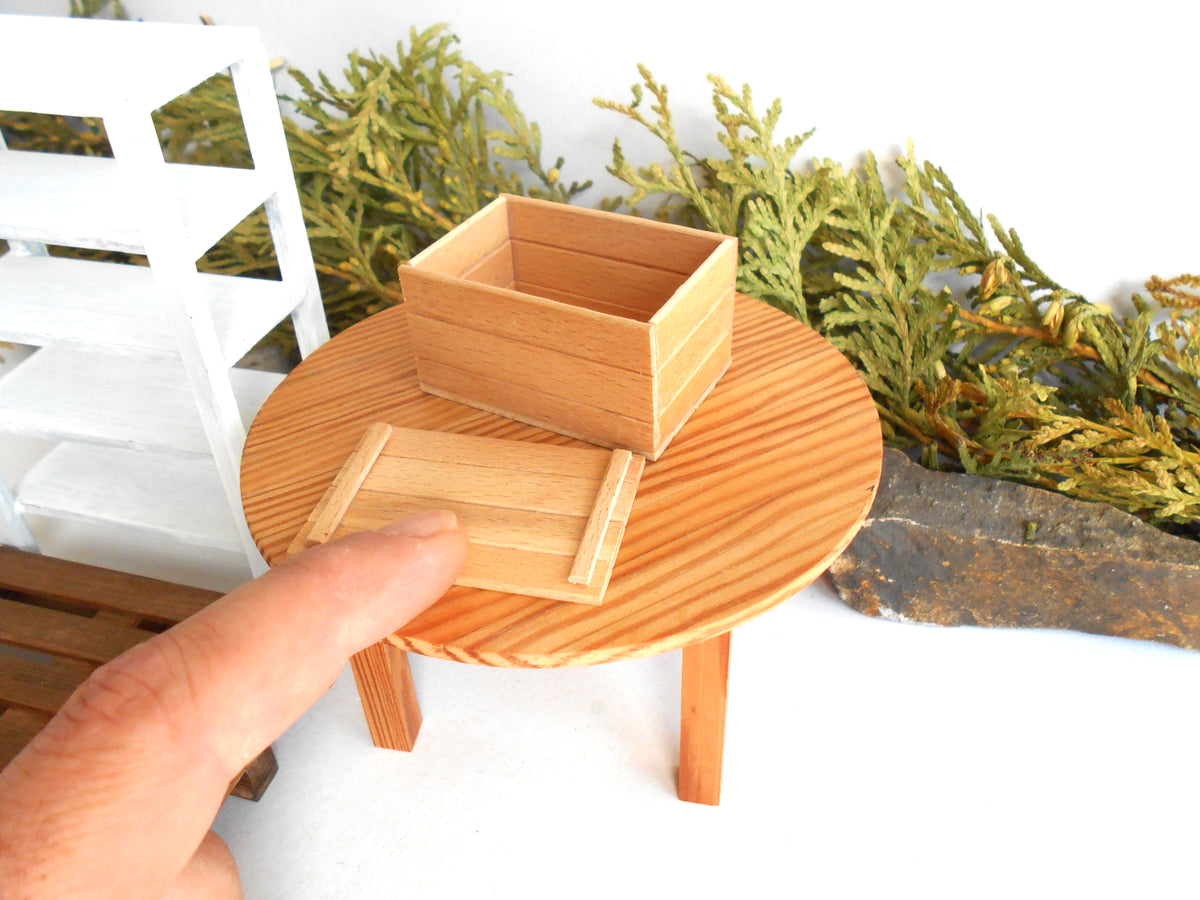 A miniature wooden storage box with its lid placed beside it on a small round wooden table, with a human finger pointing at the product. The scene includes decorative greenery and a white shelving unit in the background.