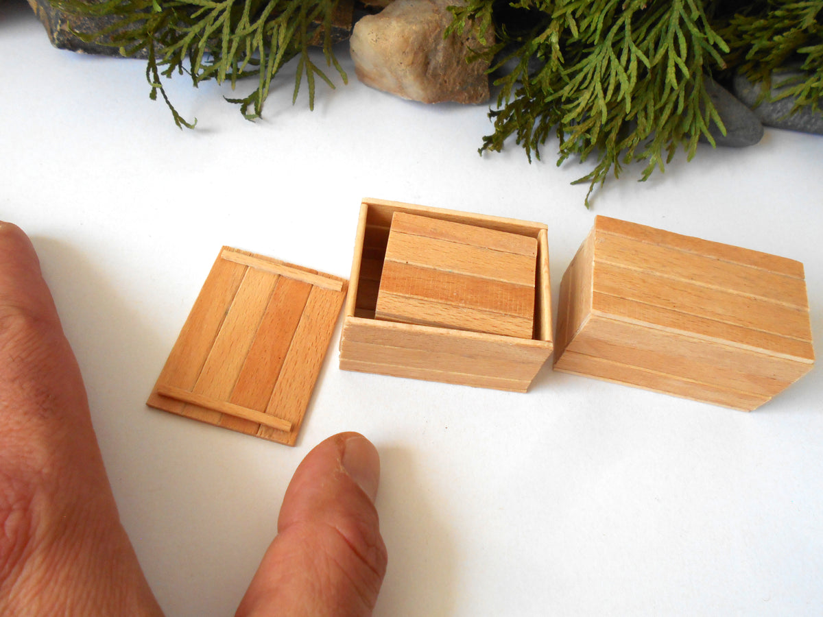 Three miniature light wood boxes, including one open box with a smaller box inside and one lid, shown on a white surface with a hand pointing at them and green foliage in the background.