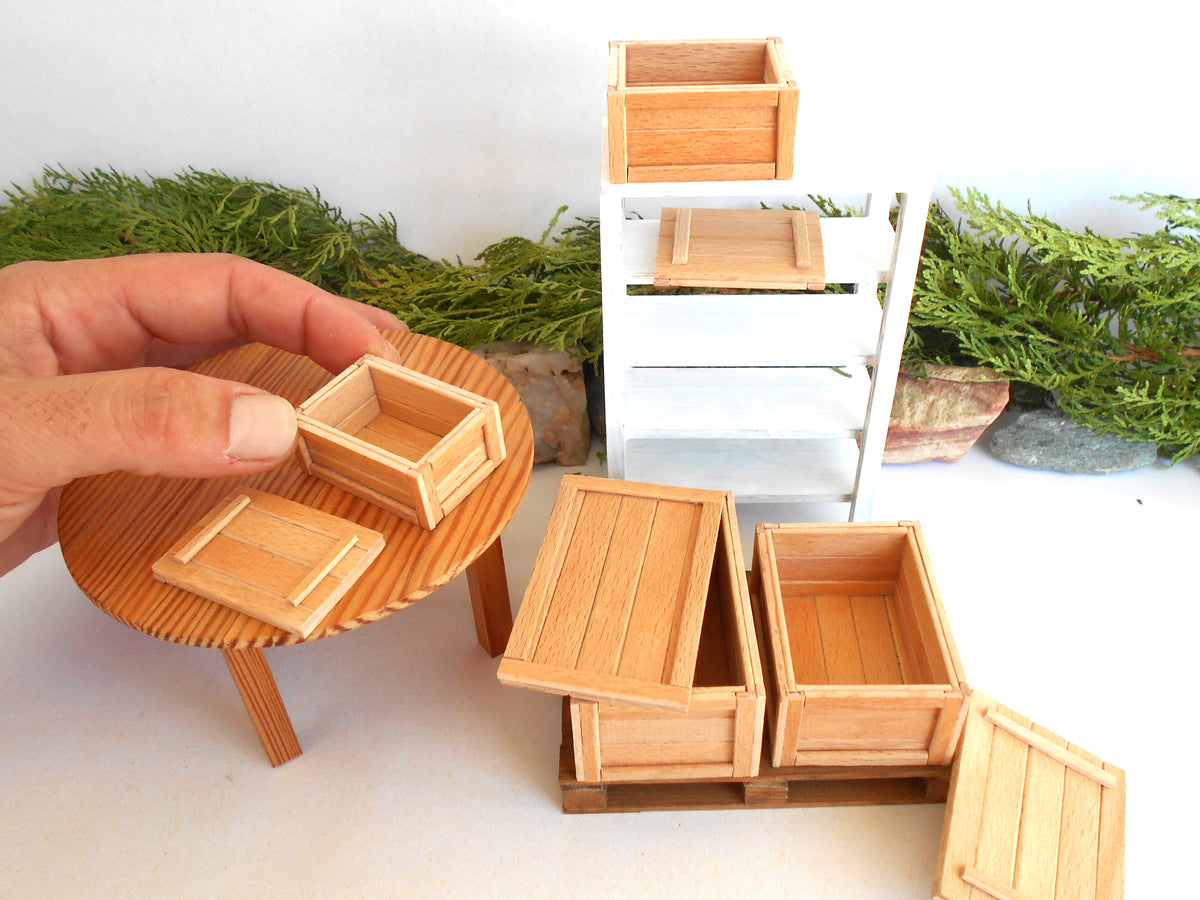 A miniature dollhouse furniture set made of light natural wood, including a small round table, storage crates, and shelving, with a hand holding one of the crates to show scale.