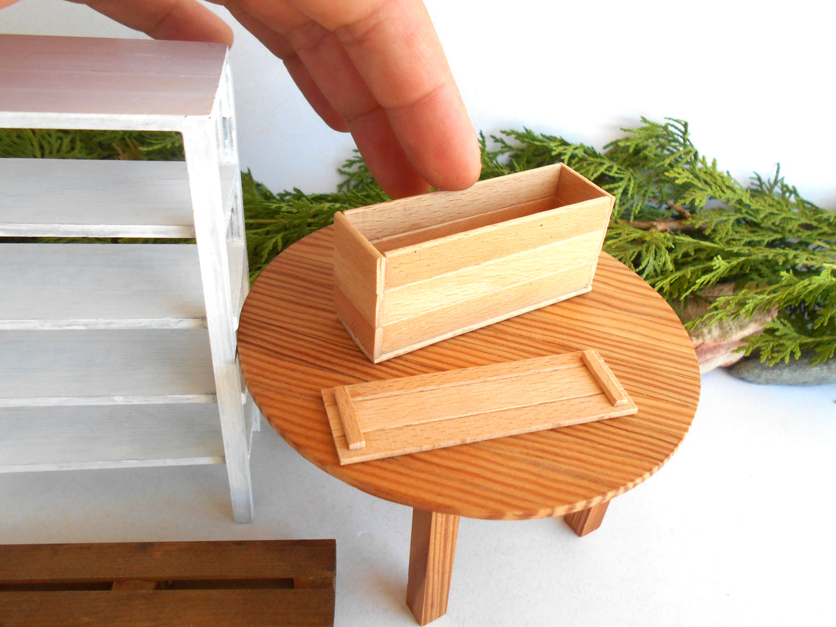 A miniature wooden planter box with its matching lid placed on a small round wooden table, with a human hand reaching toward the planter. The scene includes a white shelf on the left and green foliage in the background.
