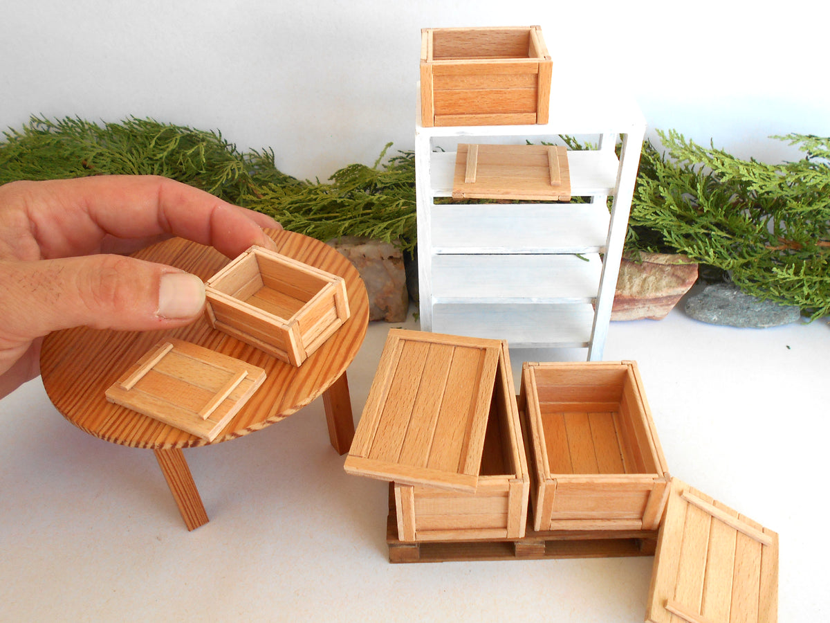 A miniature wooden storage crate set with matching lids, shown alongside a small round wooden table and a white shelving unit. A human hand is holding one of the crates on the left, and the background includes decorative green foliage against a white wall.