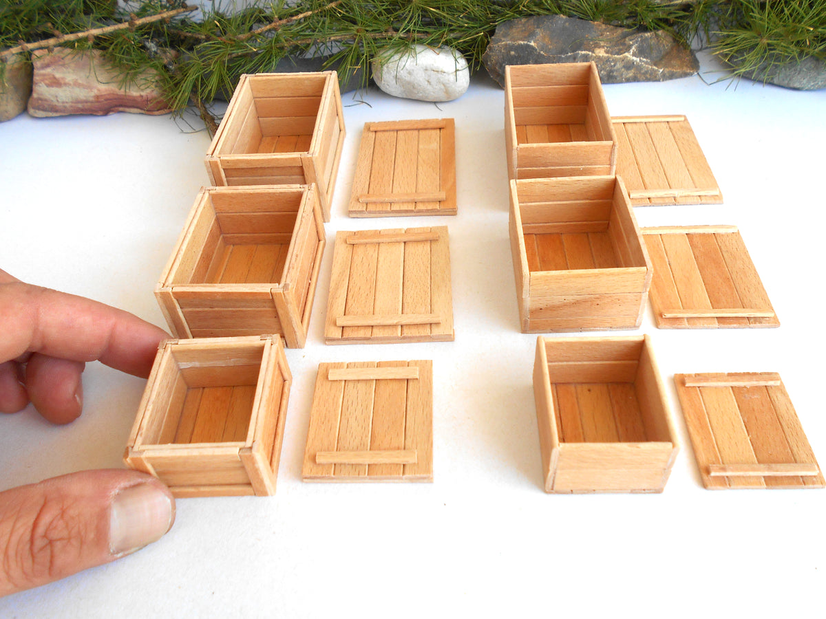 A hand is holding one of several miniature light wood crates or boxes arranged on a white surface, with small stones and greenery in the background.