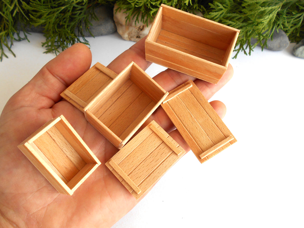 Five miniature light brown wooden boxes with a visible wood grain texture, held in a person's hand against a white background with green foliage at the top.