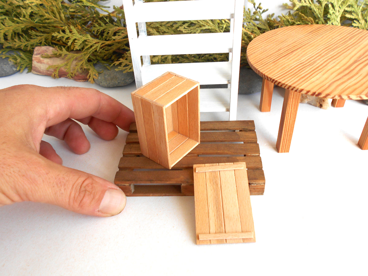 A miniature wooden crate set with a hand adjusting one of the small wooden boxes on a white surface, with a miniature white chair and round wooden table in the background.