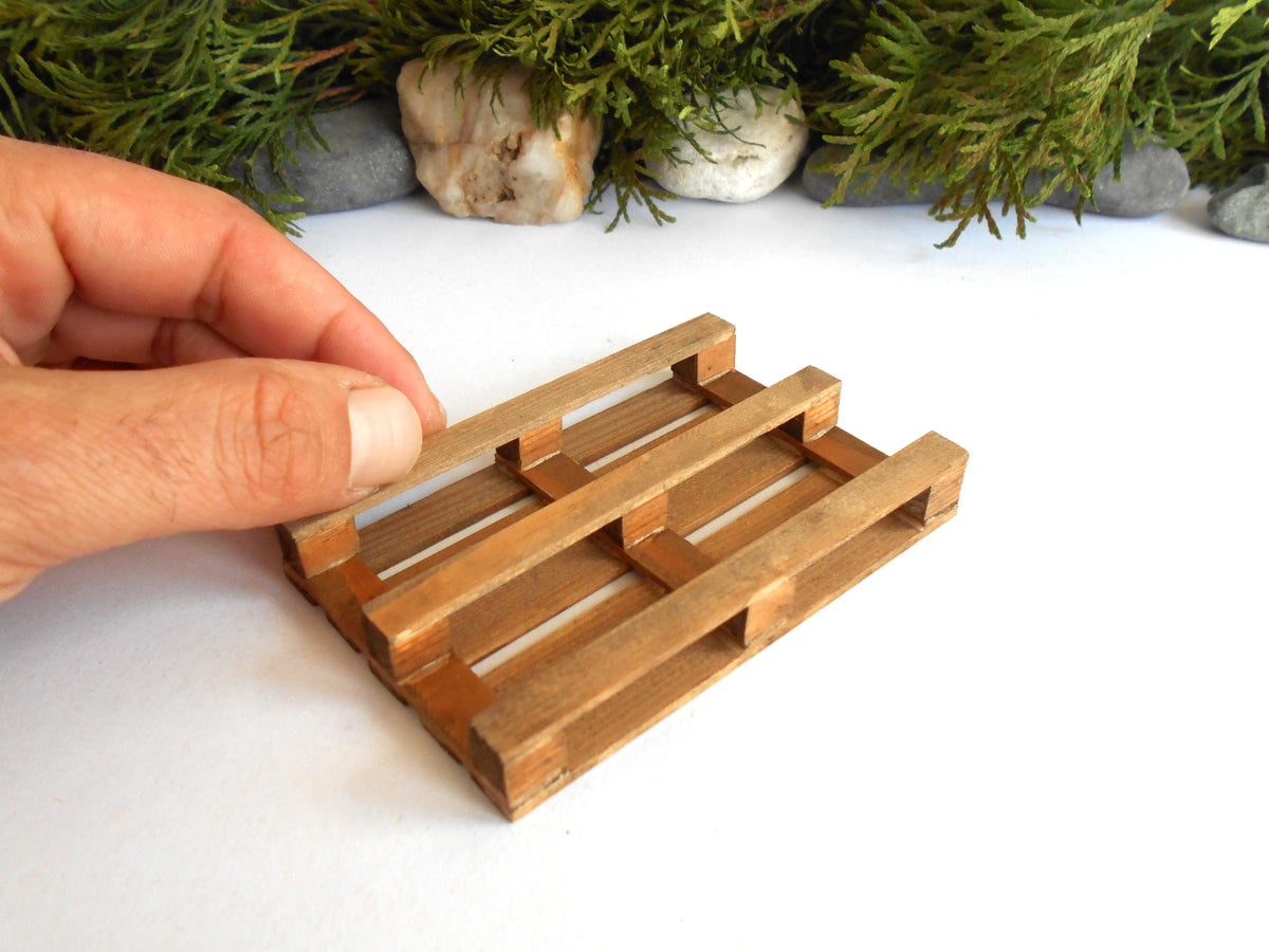 A miniature wooden pallet model being held by a hand against a white background with decorative greenery and stones at the top.