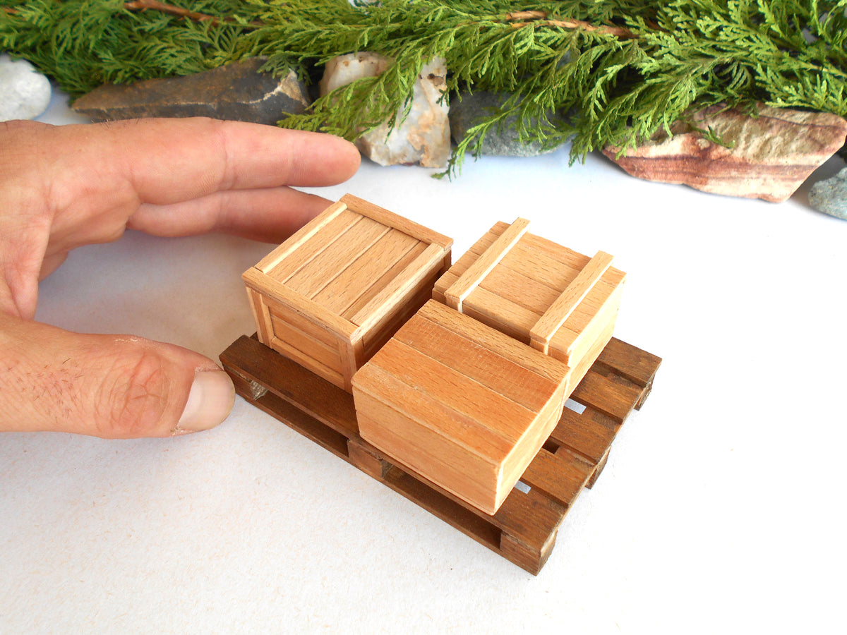 A close-up of a miniature wooden toy or model featuring two small stacked boxes on a miniature pallet, with a human hand pointing toward it for scale against a white background with decorative greenery.