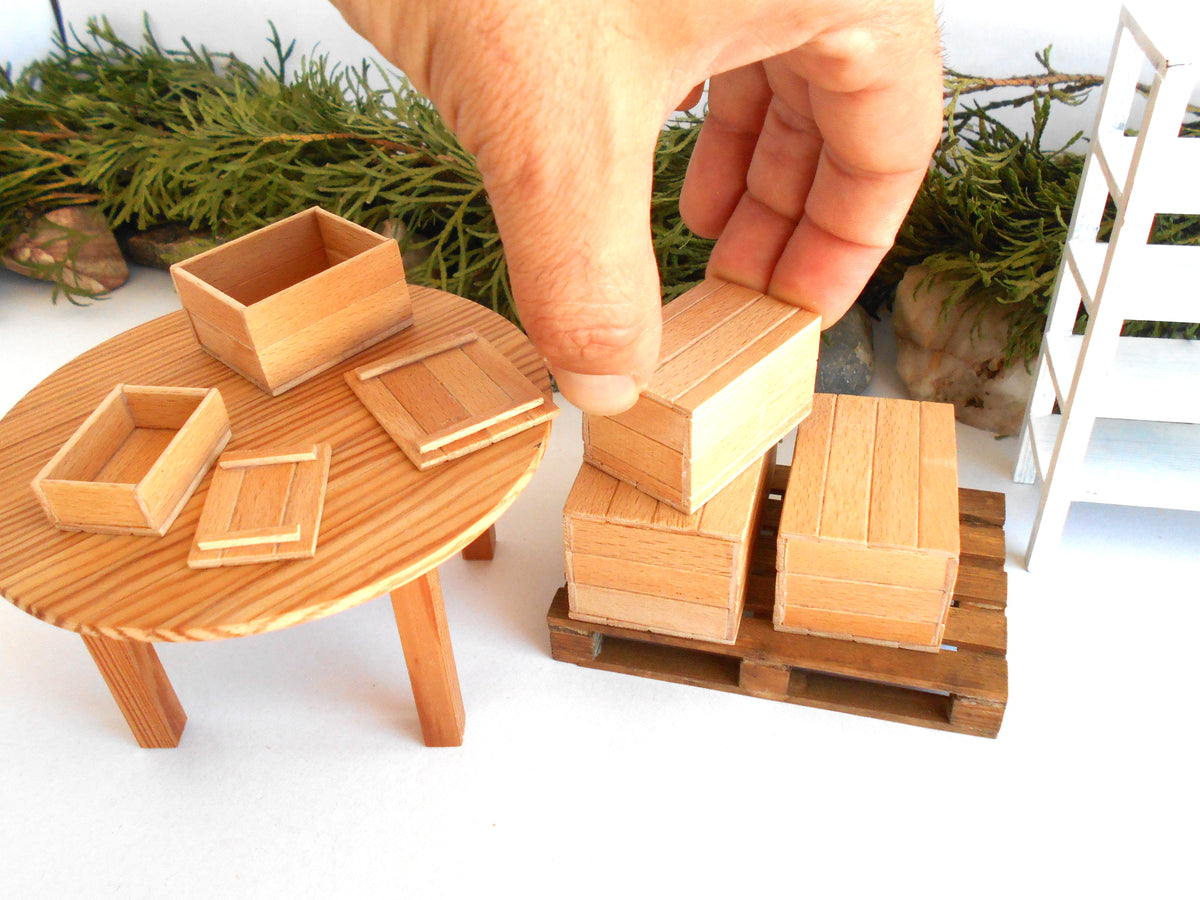 A hand is placing a small wooden box onto a miniature pallet in a detailed diorama setup. The scene includes a round wooden table with small wooden crates and boxes, green pine branches in the background, and a white miniature ladder on the right.