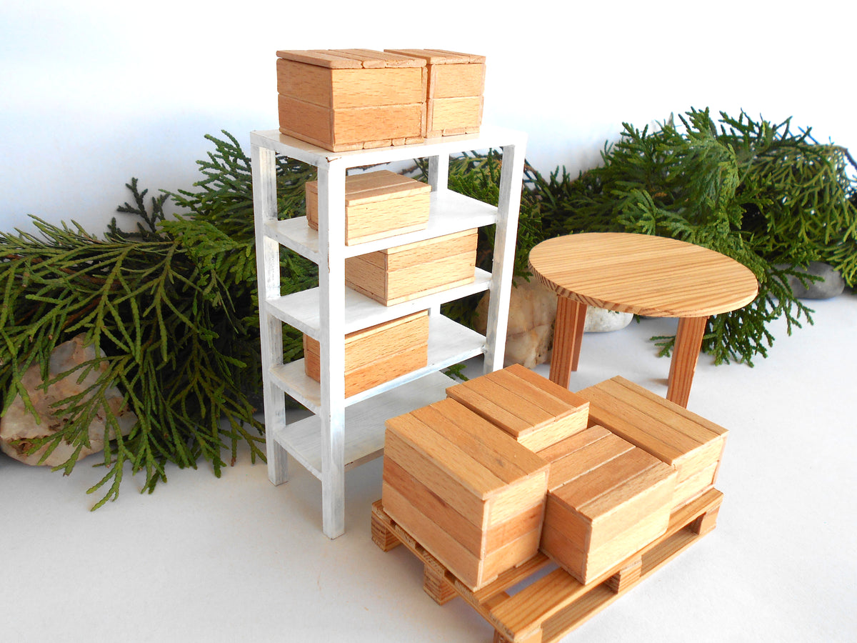 A miniature wooden toy furniture set featuring a white three-tier shelf with stacked light wood boxes, a round light wood table, and a wooden pallet with matching boxes, styled with green pine branches on a white surface.