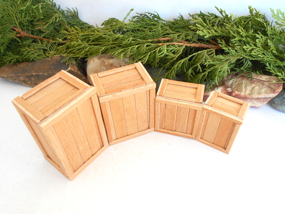 Four miniature light wood crates arranged in a curved row on a white surface, with green foliage and small stones behind them.