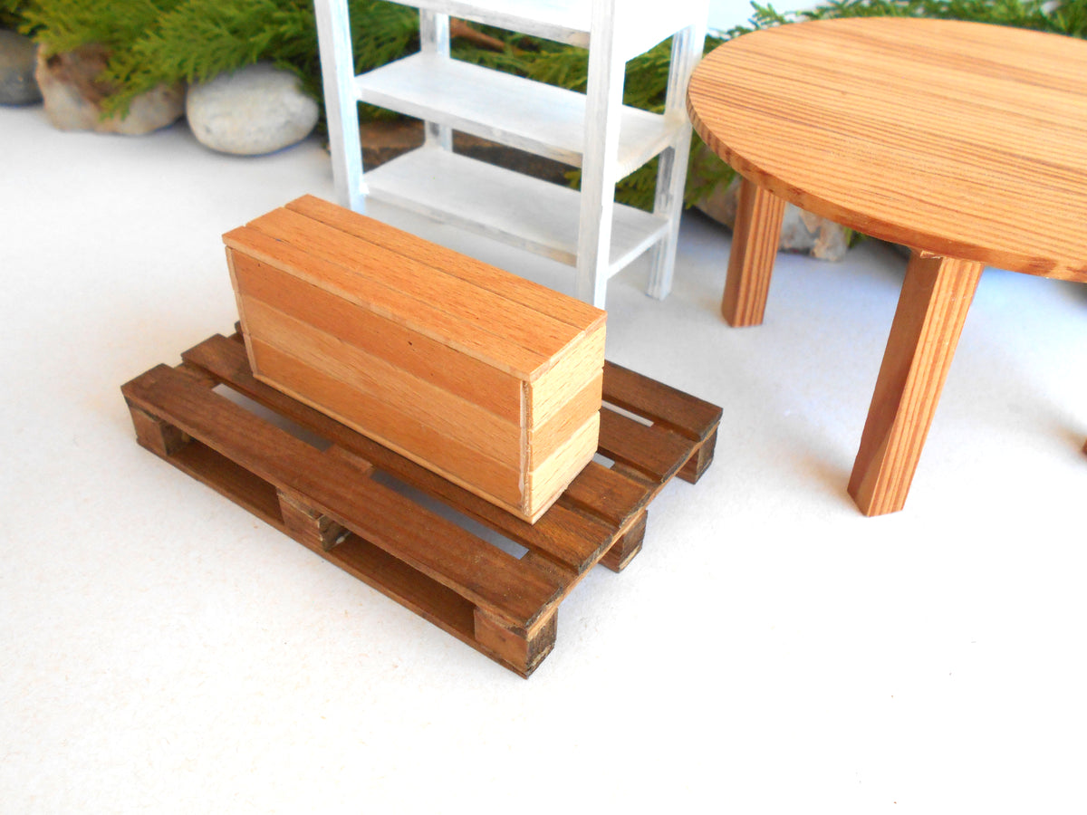 A miniature wooden box on a small wooden pallet, with a miniature white shelf and round wooden table in the background.
