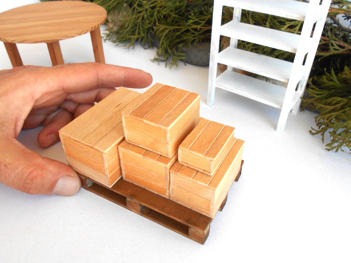 A miniature wooden toy truck carrying several small wooden cargo boxes, with a human hand holding it for scale, set against a white tabletop with miniature furniture props.