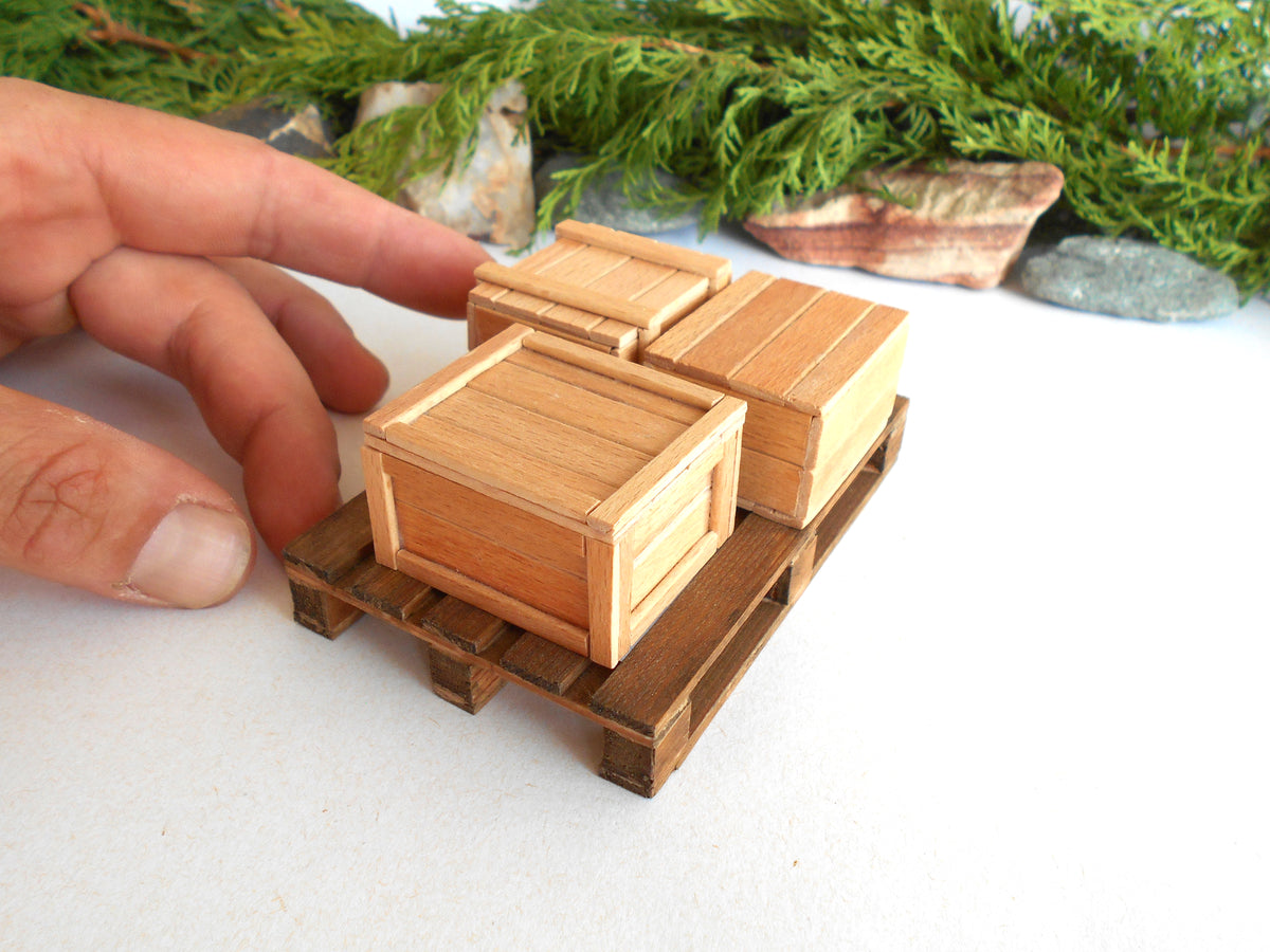 A miniature wooden crate set on a small wooden pallet, with a human hand pointing at the front crate to show scale, against a white background with decorative greenery.
