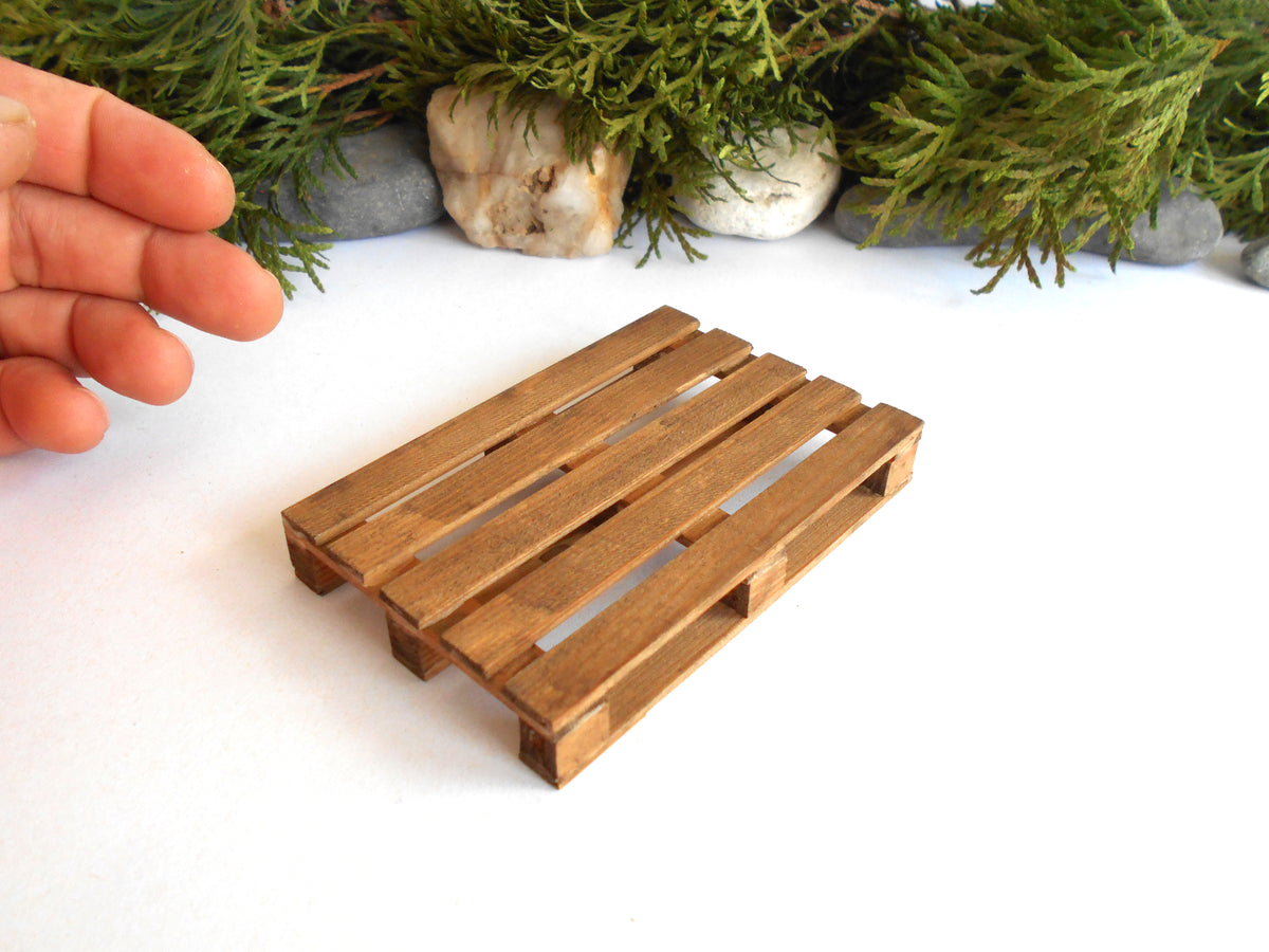 A miniature wooden pallet toy or scale model, shown on a white surface with a hand pointing near it and green foliage in the background.