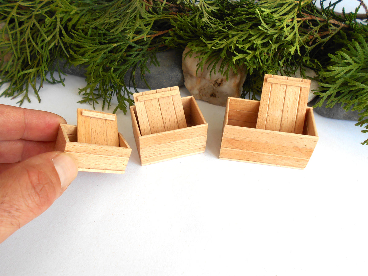 Three miniature light wood box-like dollhouse accessories displayed on a white surface, with a hand holding the smallest one for scale and green foliage in the background.