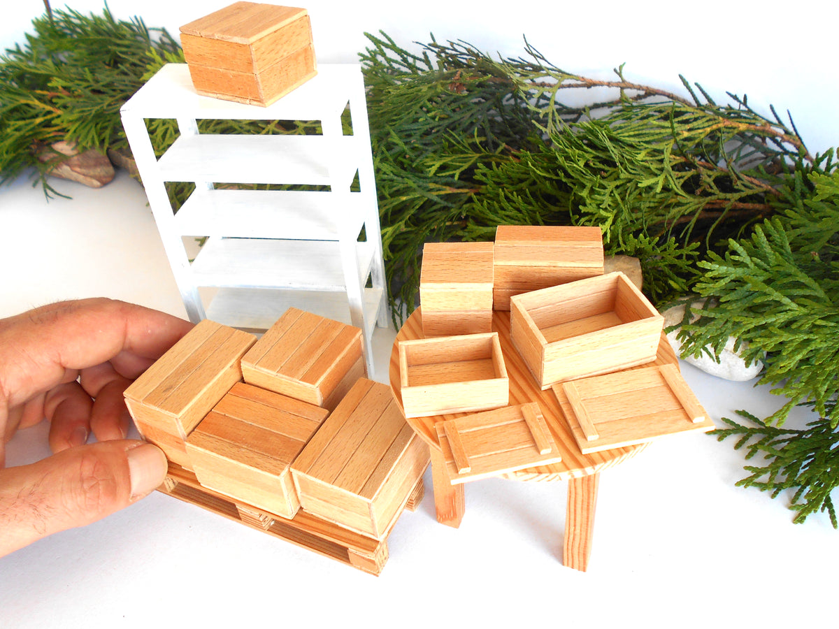 A hand holds a miniature wooden storage rack set, including a pallet-style crate stack and a small table with open boxes, alongside a white shelving unit and green pine branches on a white background.