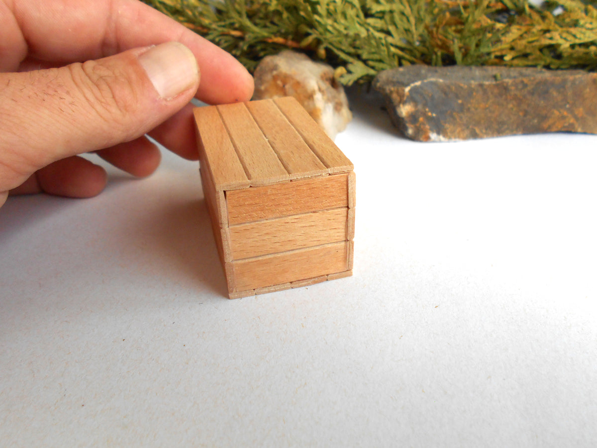 A hand holding a miniature wooden box with three drawers, shown in close-up against a white surface with blurred natural background elements.