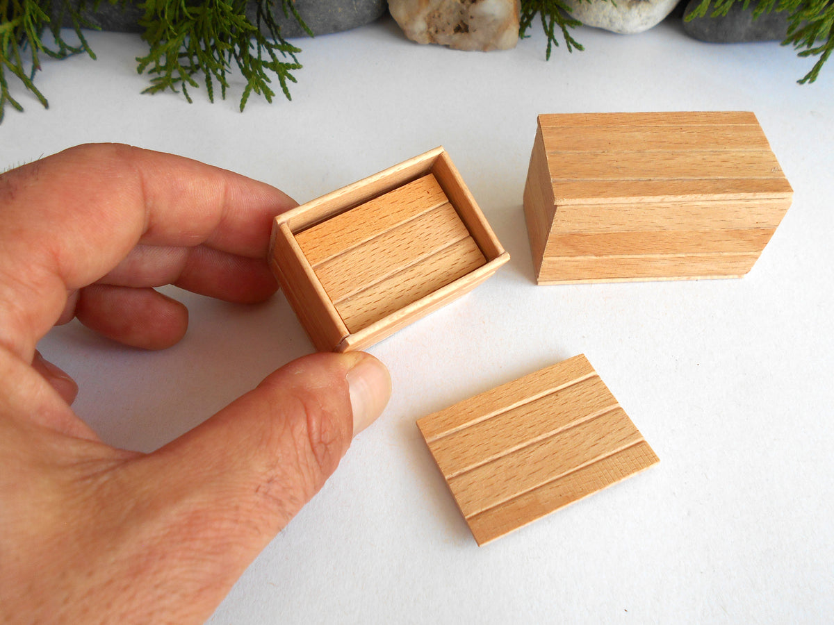 A hand holds a small rectangular wooden box with a matching lid placed beside it on a white surface. A second closed wooden box sits in the background, with blurred greenery and stones at the top edge.