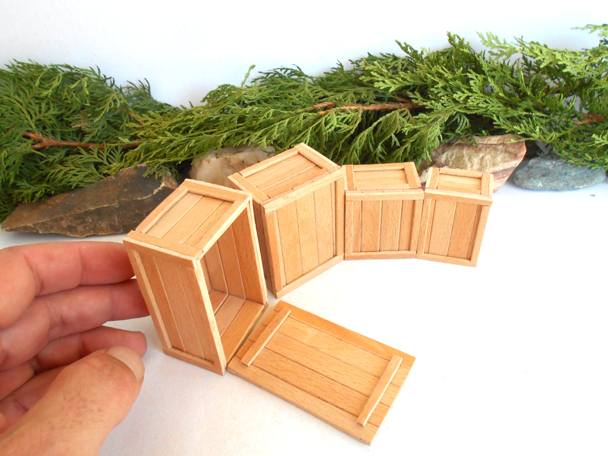 A hand holding a miniature wooden crate next to several other small wooden crates, with green foliage and rocks in the background.