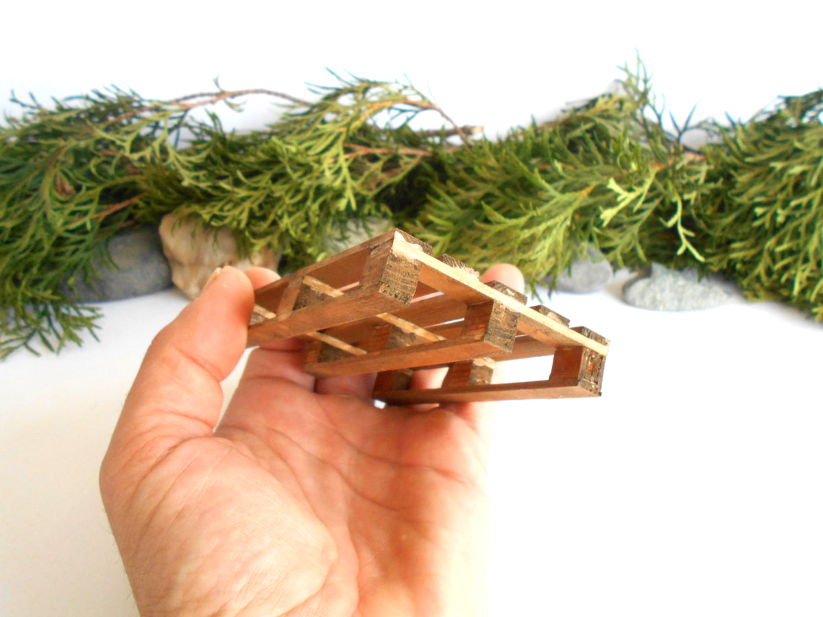 A hand holding a miniature wooden pallet structure, with green garland and small stones in the background.