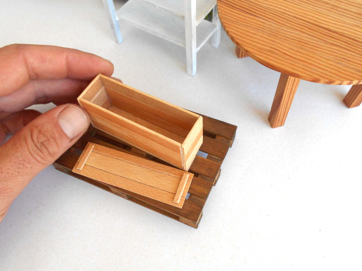 A hand holding a small rectangular wooden box over a miniature wooden base, with a miniature round table and shelf in the background.