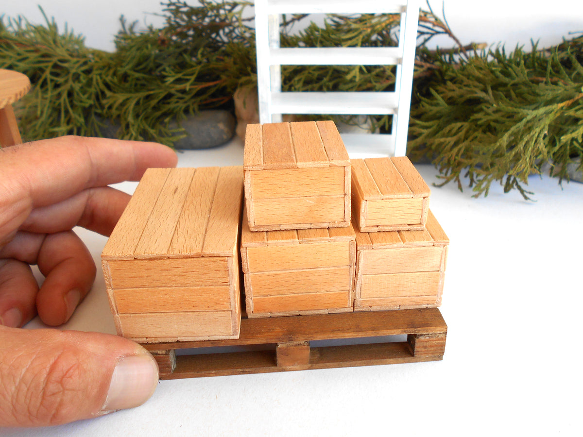 A hand holding a miniature wooden pallet with several small stacked wooden crates, shown in a close-up product photo with a white surface and blurred greenery in the background.