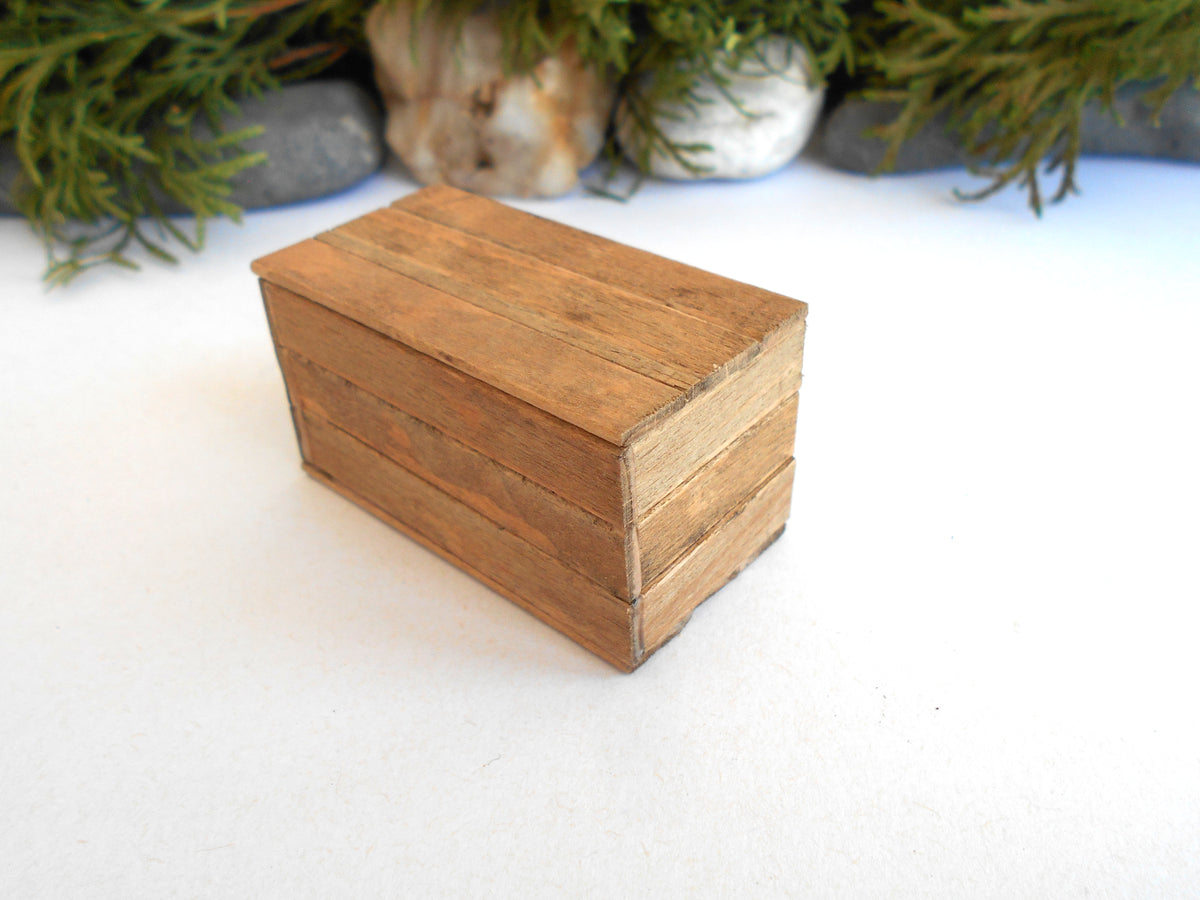 A miniature wooden box with a rustic, weathered finish, photographed on a white surface with blurred green foliage in the background.
