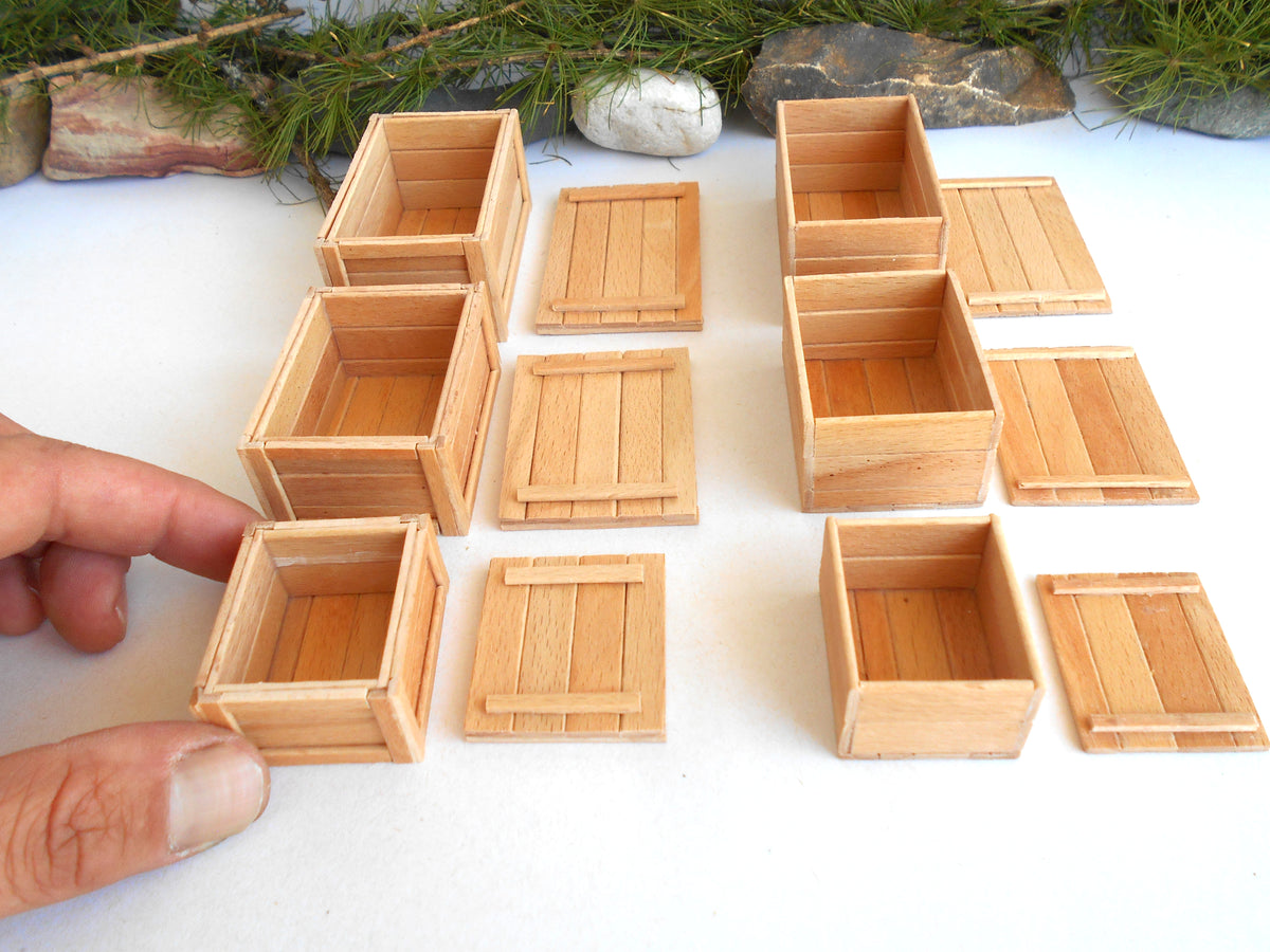 A set of miniature light wood crates and lids, with a hand holding one of the small crates on a white surface with decorative stones and pine branches in the background.
