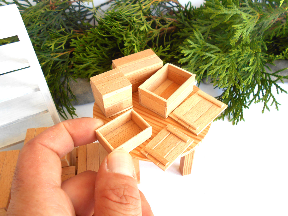 A hand holding a miniature wooden table and storage crate set, with additional matching wooden boxes arranged on a white surface and green foliage in the background.