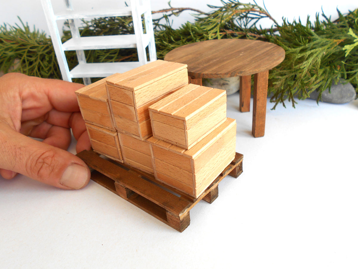 A miniature wooden building set for dollhouses, featuring a stack of three small wooden houses on a wooden pallet, with a hand holding it in place. The set includes a small round wooden table and a white ladder in the background.