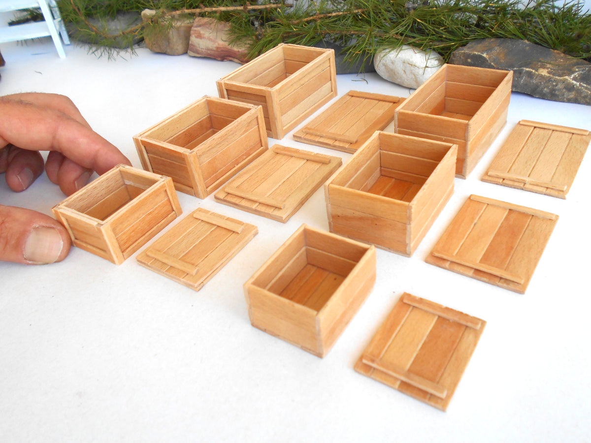 A set of miniature light brown wooden boxes and lids, shown with a person's hand holding one of the small boxes to demonstrate scale, on a white surface with a decorative background.