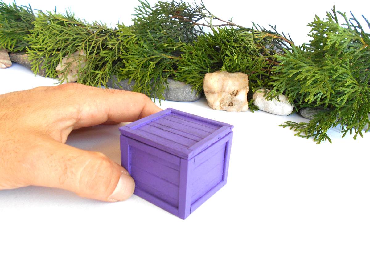 A miniature purple wooden crate box being held by a human hand, with green foliage and small stones in the background on a white surface.