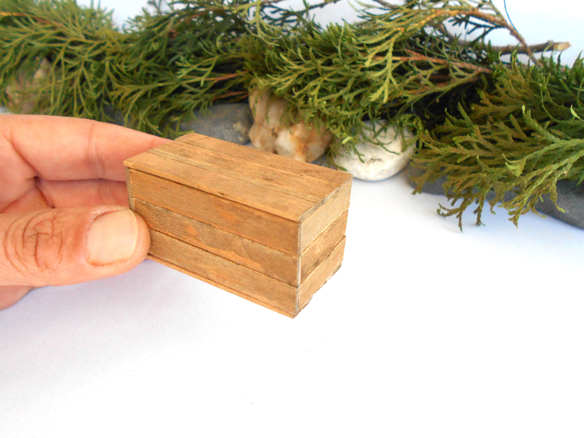 A hand holding a small, rectangular wooden box with three stacked layers, set against a white background with green foliage and stones in the blurred background.