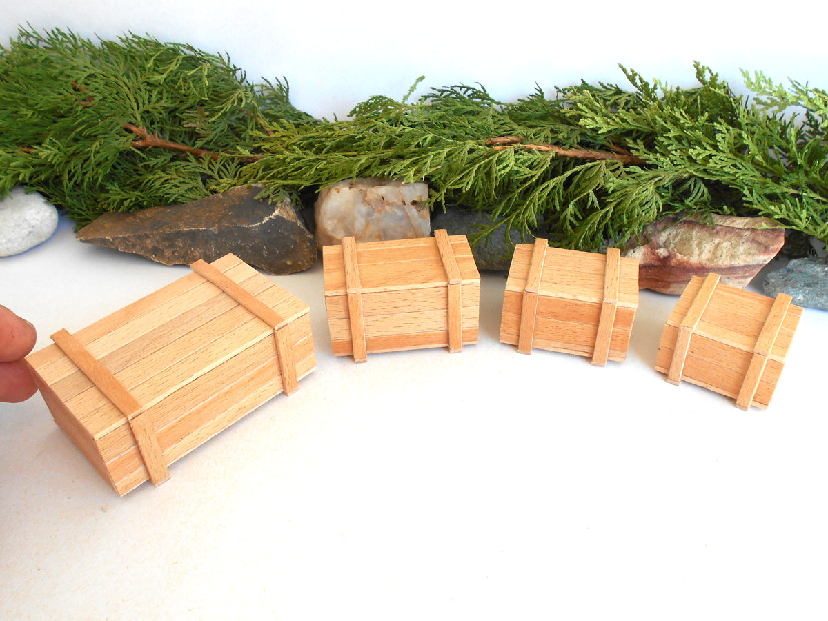 Four miniature light wood toy or model crates arranged on a white surface with green foliage and stones in the background. A human finger is visible at the far left edge.