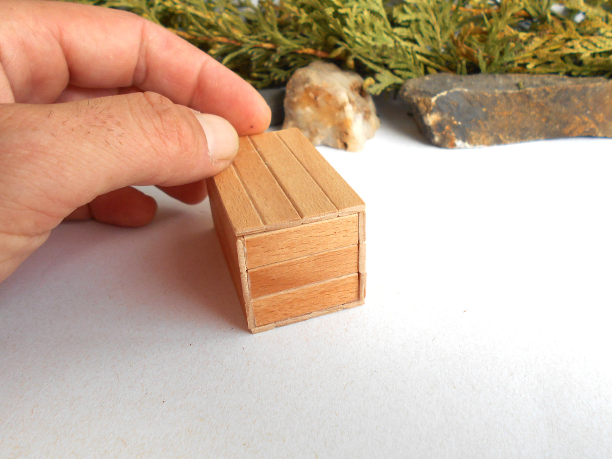 A hand holding a miniature wooden box with a slatted top, shown close-up on a white surface with small decorative props in the background.