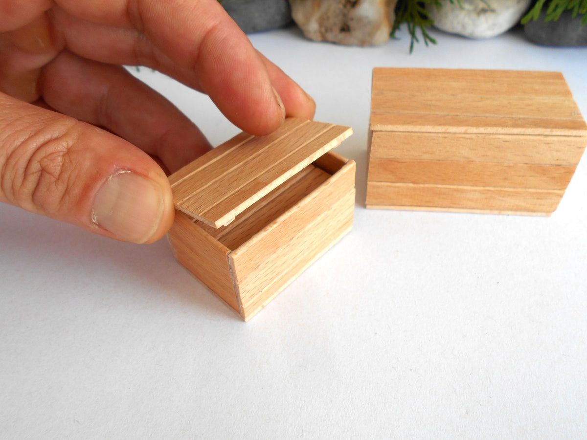 A close-up of a hand holding a small wooden box with its lid slightly open, next to another closed matching wooden box on a white surface.