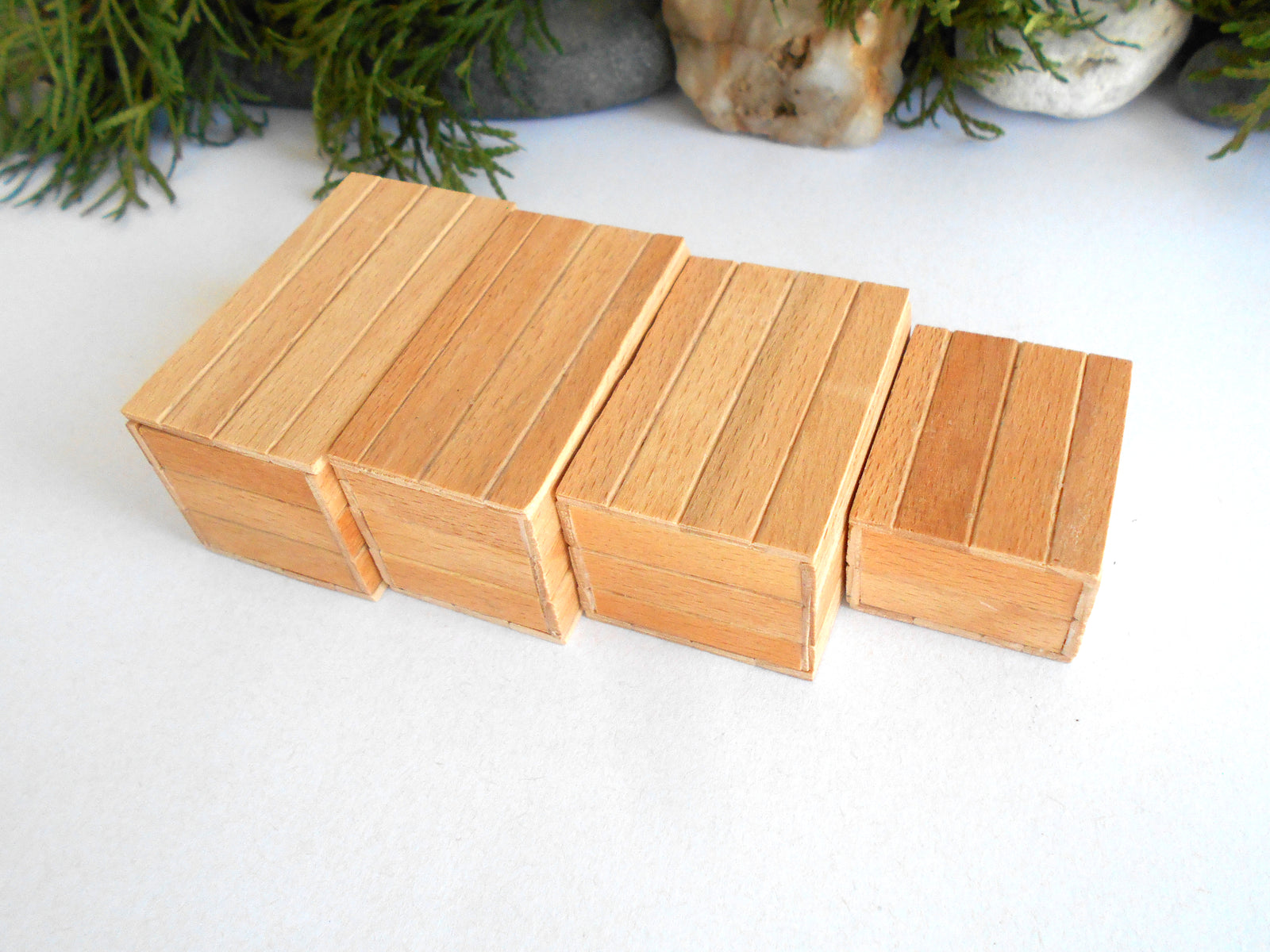 Four small rectangular mini wooden boxes arranged in a staggered row on a white surface, with decorative greenery and stones blurred in the background.