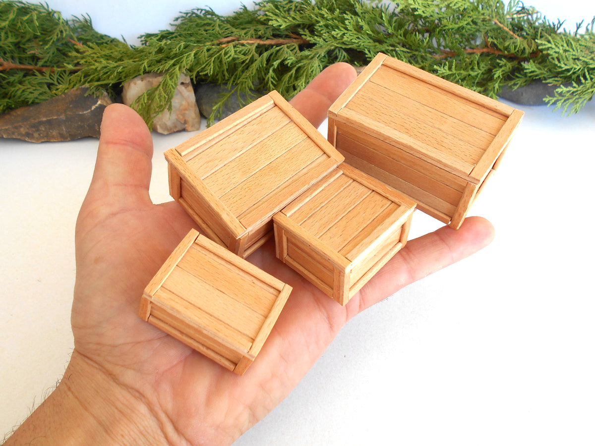 Four miniature light wood crates of different sizes held in a person's hand, with green pine branches in the background.