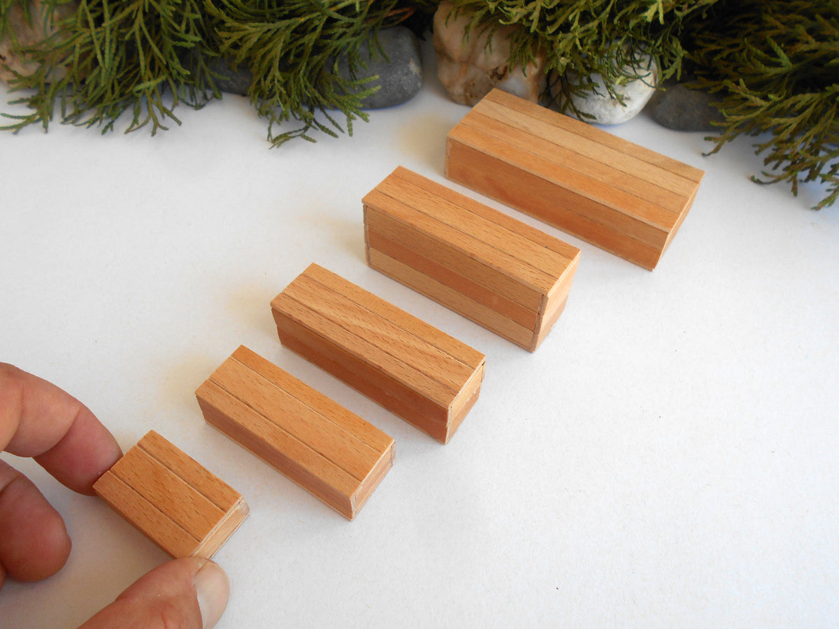 Five small rectangular wooden miniature boxes of varying sizes arranged diagonally on a white surface, with a hand holding one of the blocks at the bottom left. Green foliage and small stones decorate the background.